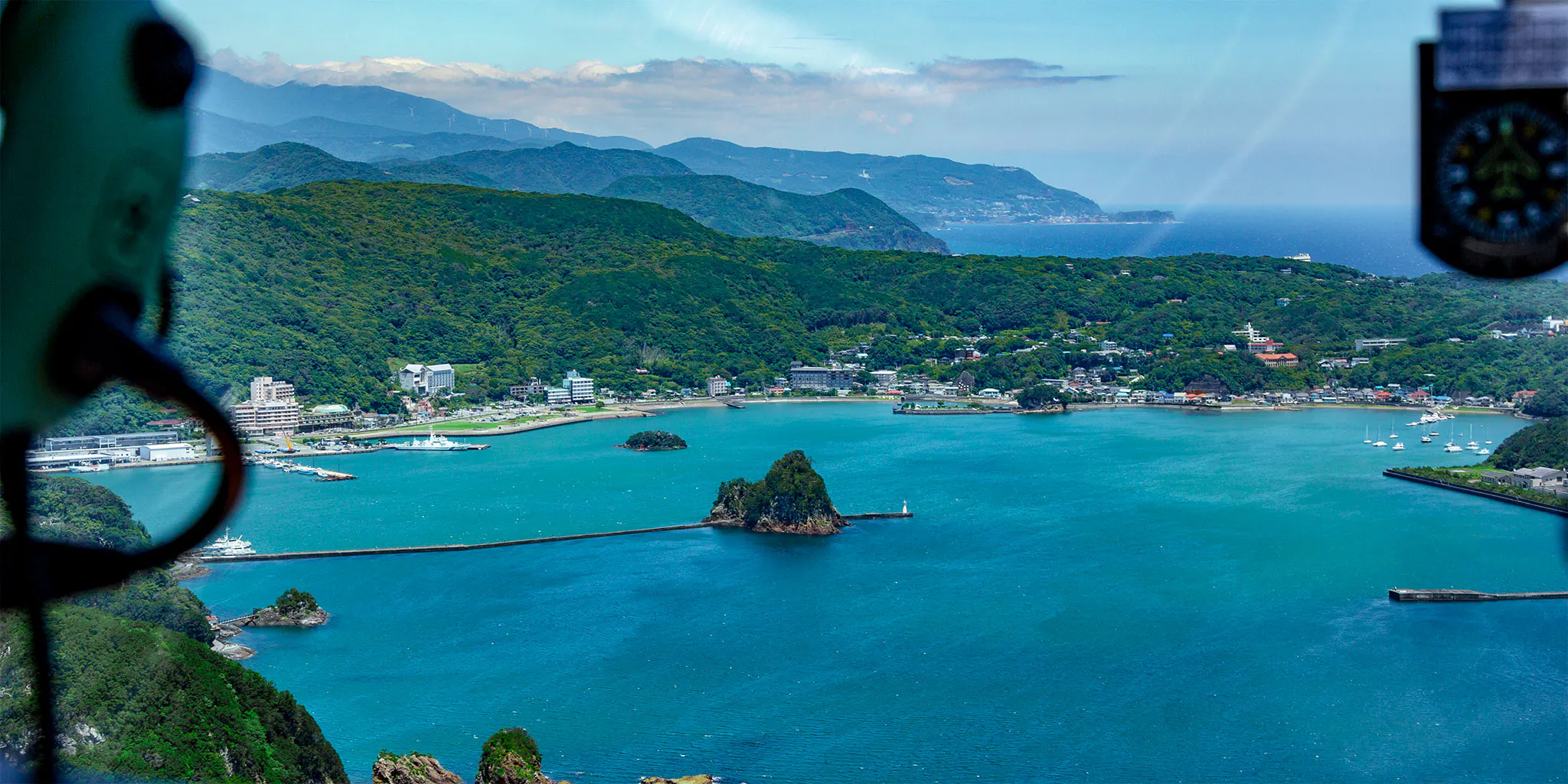 View of Shimoda Bay through the helicopter window, showing a small island linked by a breakwater and the harbor town