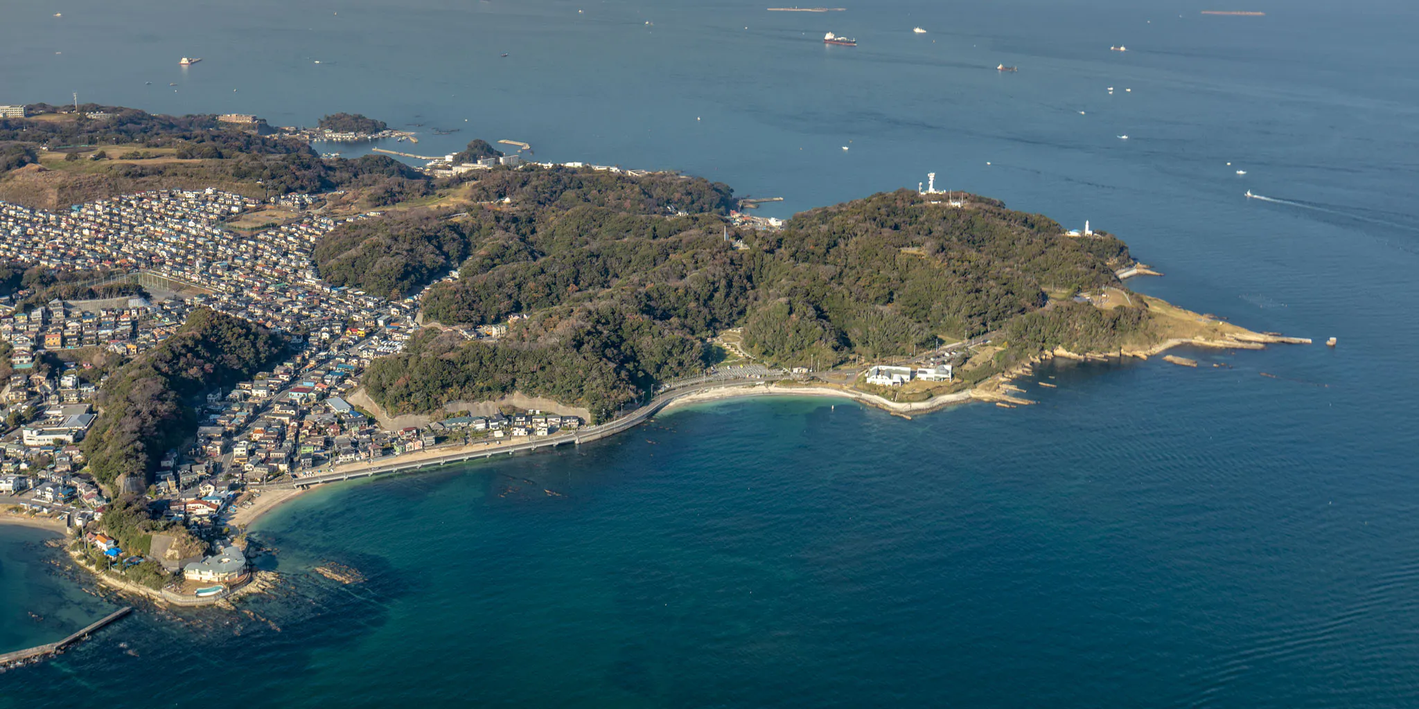 Aerial view overlooking a lighthouse-topped headland, a cove beach, and a hillside residential area