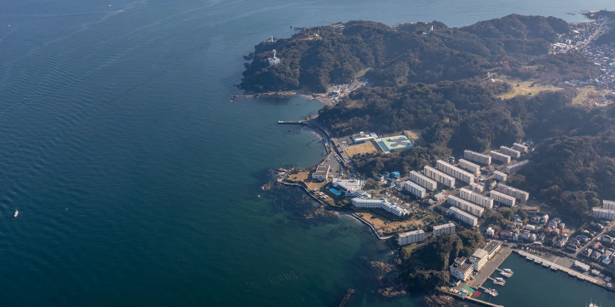 Aerial view of a poolside resort hotel and apartment blocks nestled beneath a lighthouse-capped headland