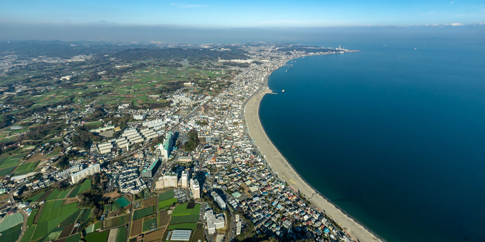 Aerial view of Miura Peninsula farmland and a shoreline village, with an industrial cape visible in the distance