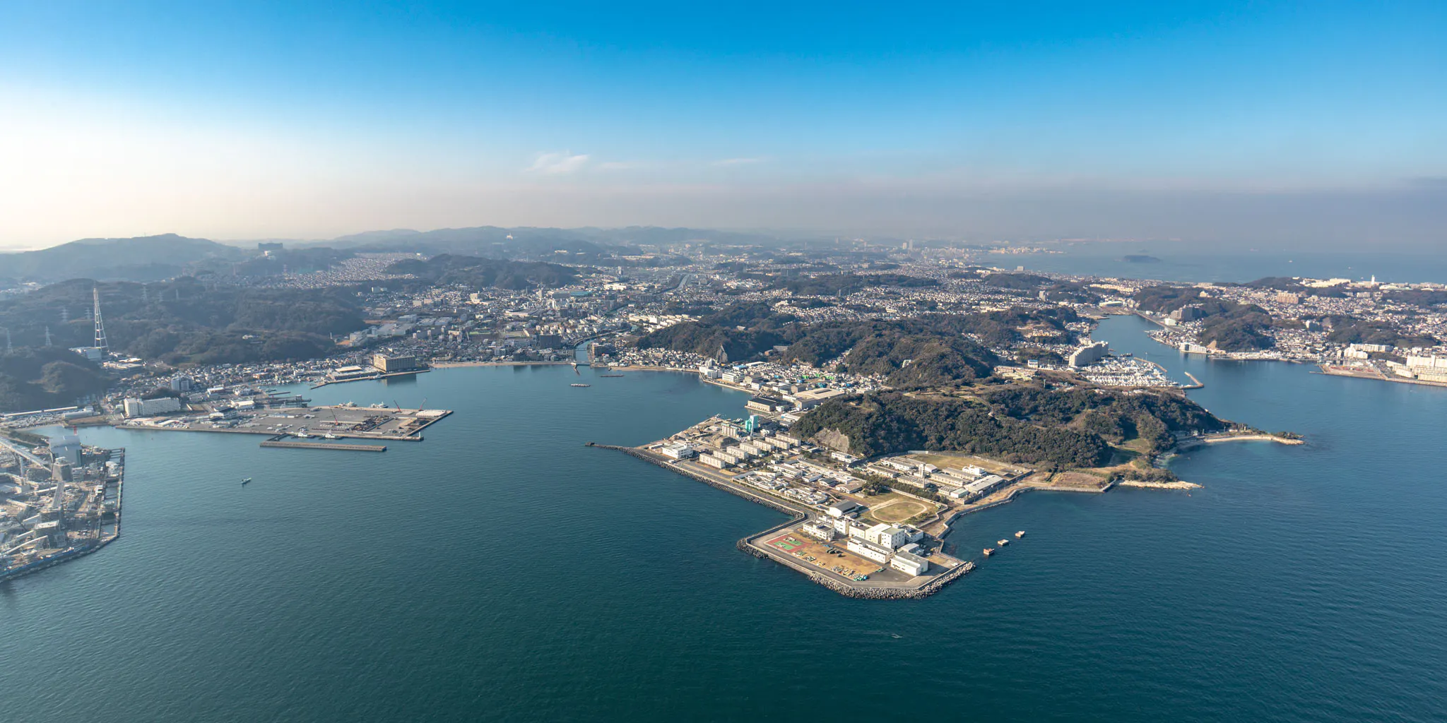 Aerial view of a cape lined with port facilities and the town of Yokosuka wrapped around the inlet