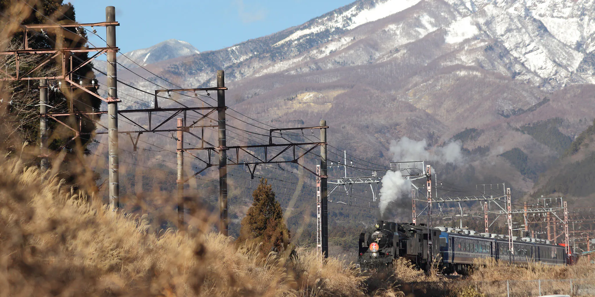 A steam train releasing white smoke, running with snow-capped mountains in the background
