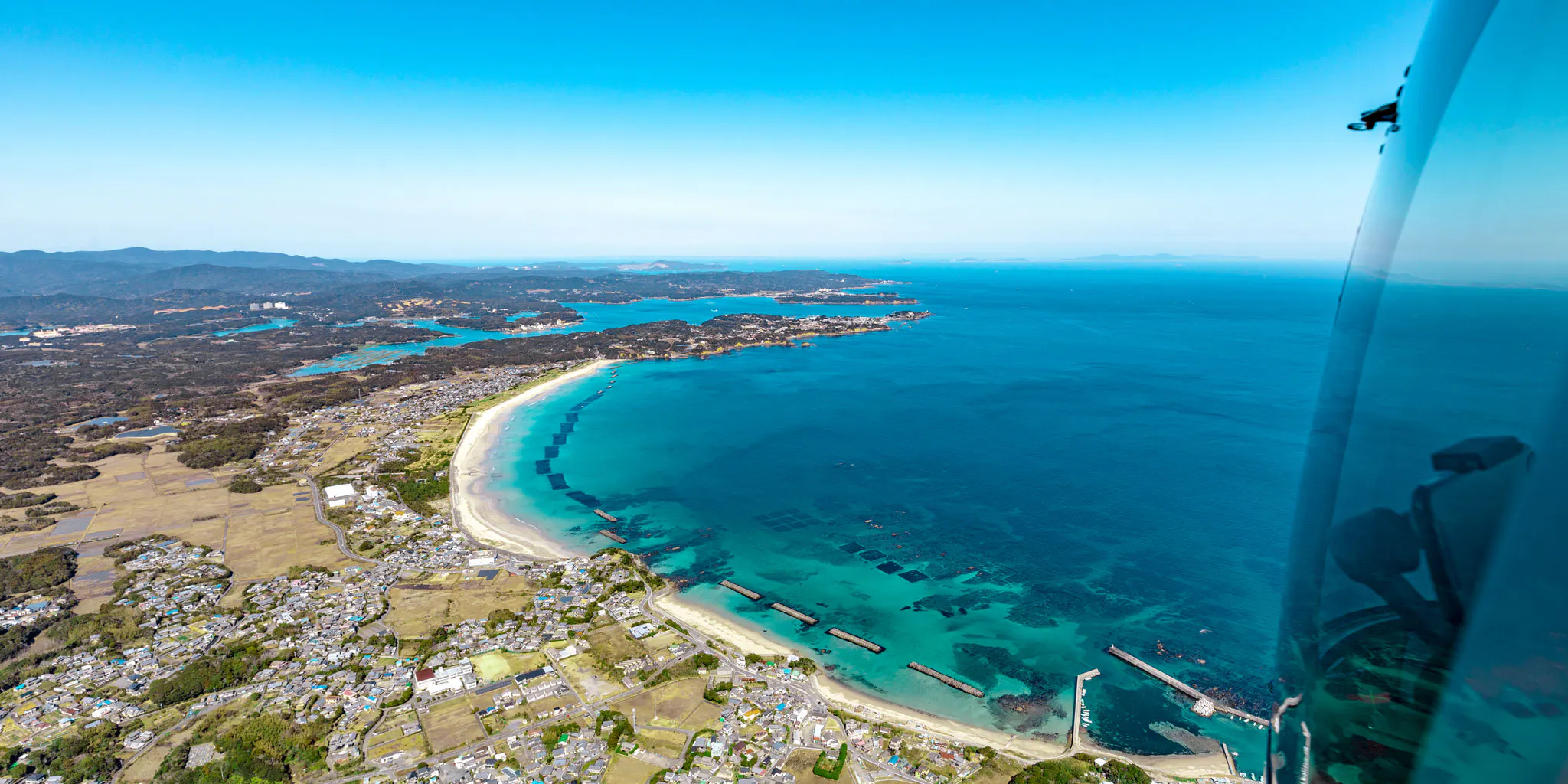 View from the helicopter cabin of a cove with aquaculture rafts and a white sand beach