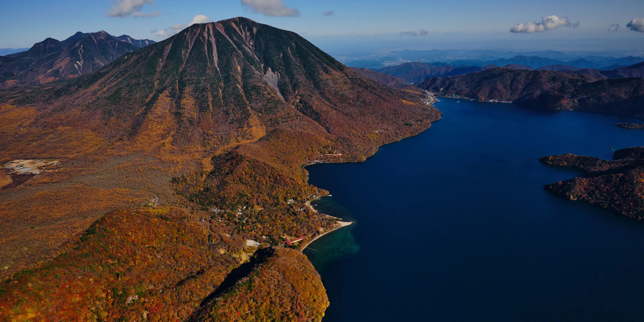 Close aerial view of the foot of Mt. Nantai and a lakeside lodge on Lake Chuzenji amid autumn foliage