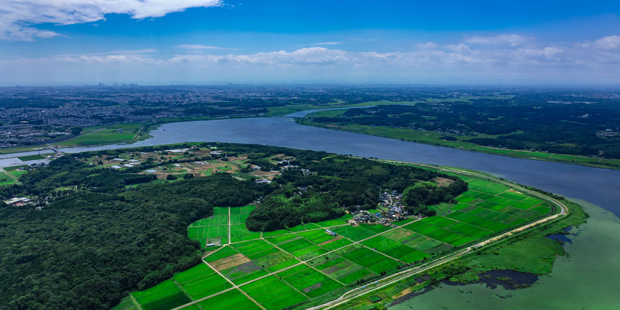 大きく蛇行する川沿いに広がる緑の農地と、小さな森と集落を上空から捉えた写真。左手の地平線には遠くの町並みが見える。