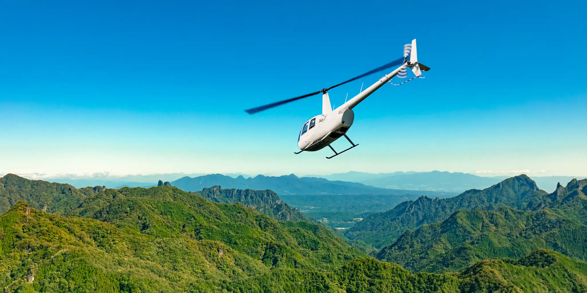 Un hélicoptère AirX survolant les montagnes verdoyantes et les pics rocheux escarpés du plateau de Chojahara, dans la chaîne du Kuju.
