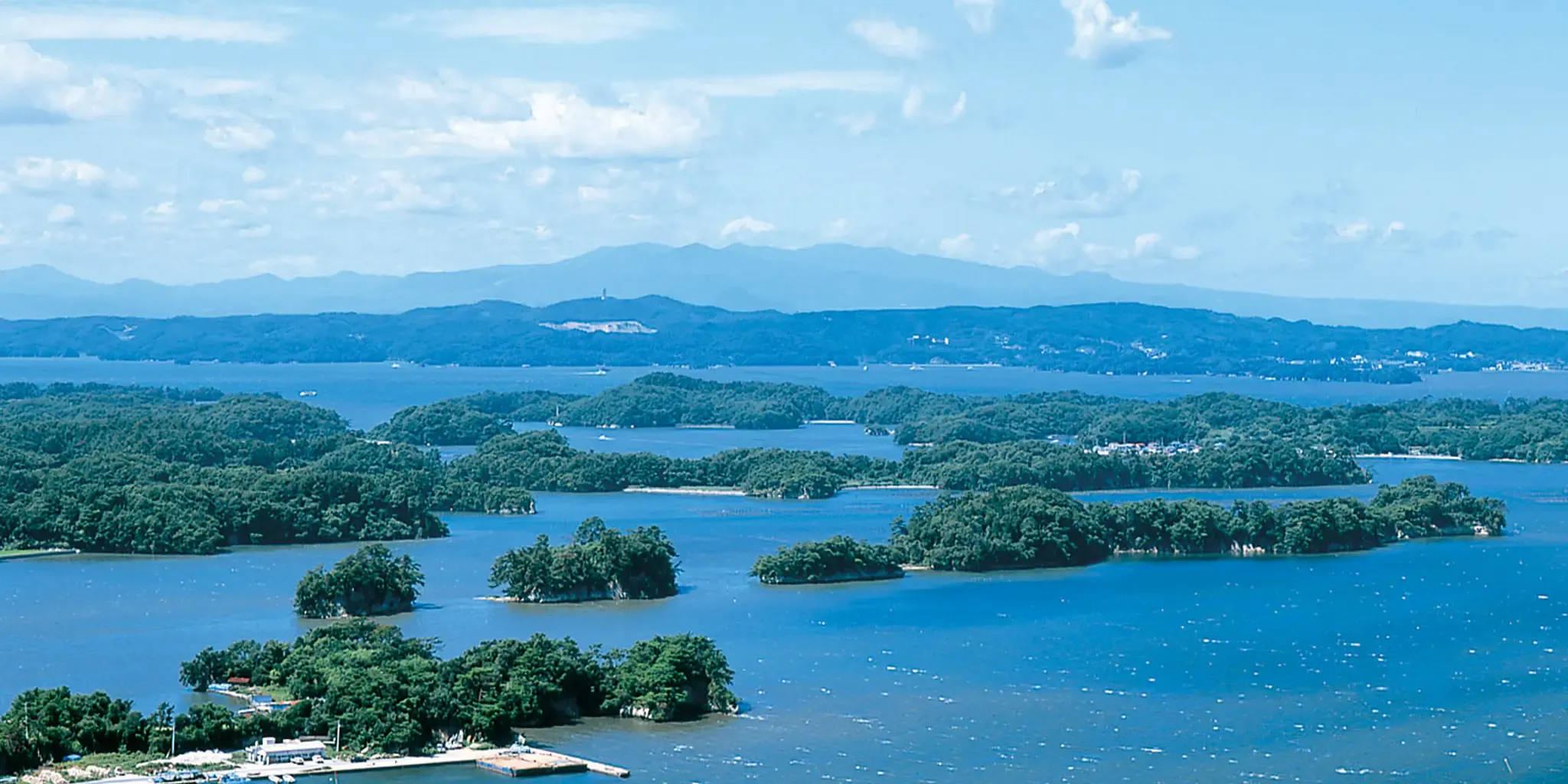 Vue panoramique sur la baie de Matsushima depuis un point de vue en hauteur, avec d'innombrables îlots couverts de pins parsemant l'eau et des montagnes embrumées à l'arrière-plan.
